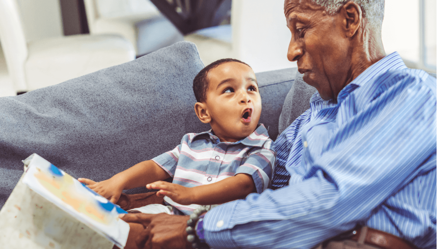 Adorable toddler with his grandfather relaxing at home reading a book together