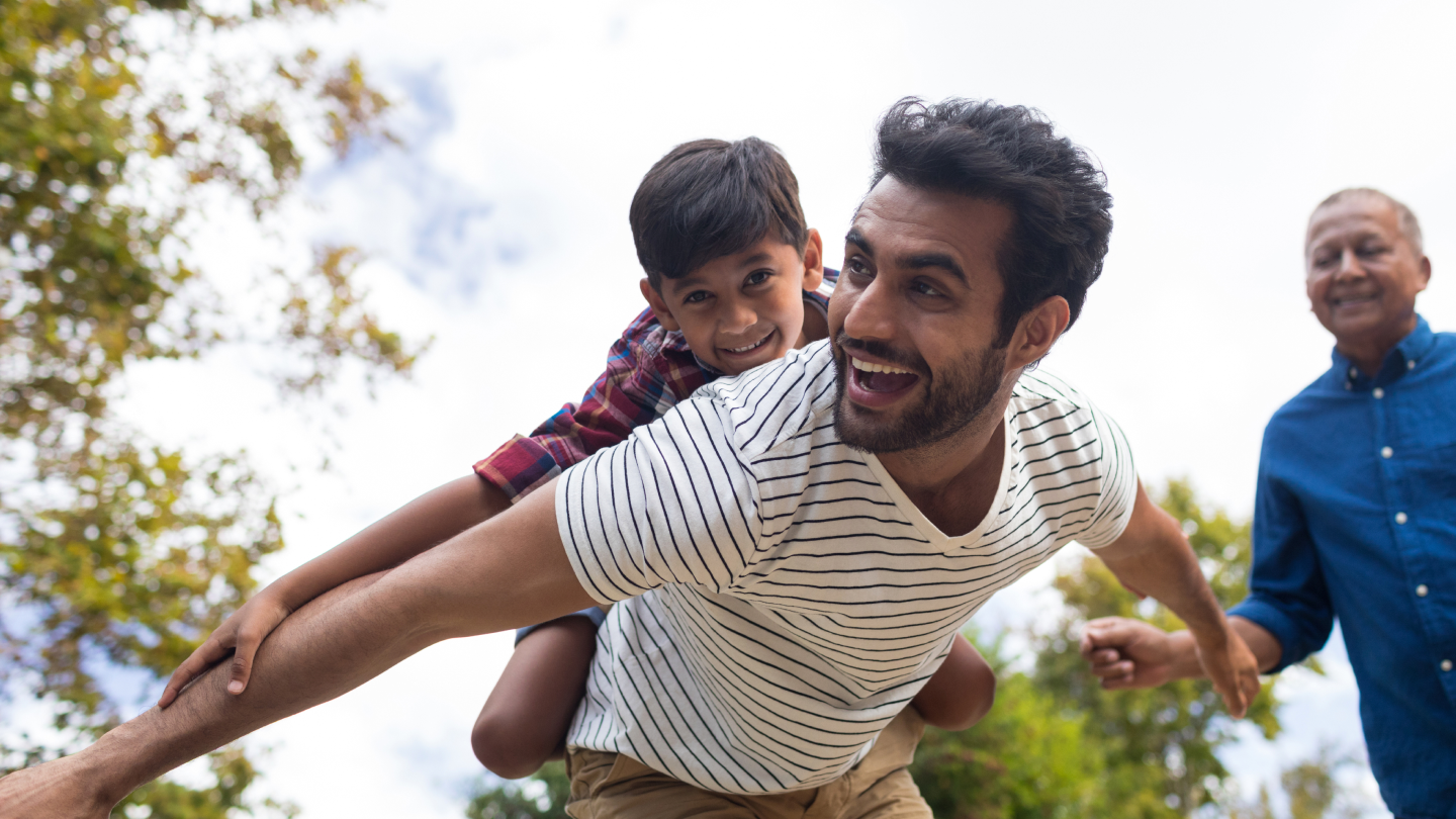 Happy grandfather looking at man giving piggy backing to son with arms oustretched in yard